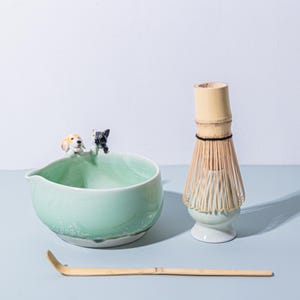 Könnte beinhalten: Eine hellgrüne Matcha-Schale aus Keramik mit einer Hunde- und Katzenfigur, einem Bambusbesen und einem Bambuslöffel. Die Schale hat einen Ausguss und einen weißen Rand. Der Schneebesen ist aus Bambus und hat einen hellgrünen Sockel.