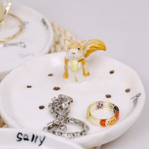 Puede incluir: Plato de cer&aacute;mica blanco para anillos con una figurita de ardilla sosteniendo una nuez amarilla. El plato tiene puntos marrones y las palabras "Sally" y "Pear" escritas en &eacute;l. Hay anillos y un collar en el plato.