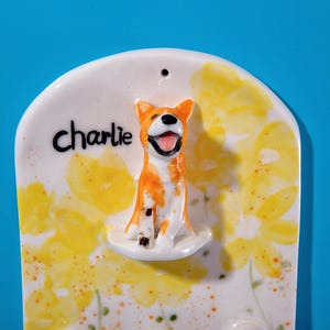 May include: A ceramic wall hanging featuring a smiling orange and white dog figurine. The dog is perched on a white base, with the name "Charlie" written above. The background is decorated with yellow floral patterns.