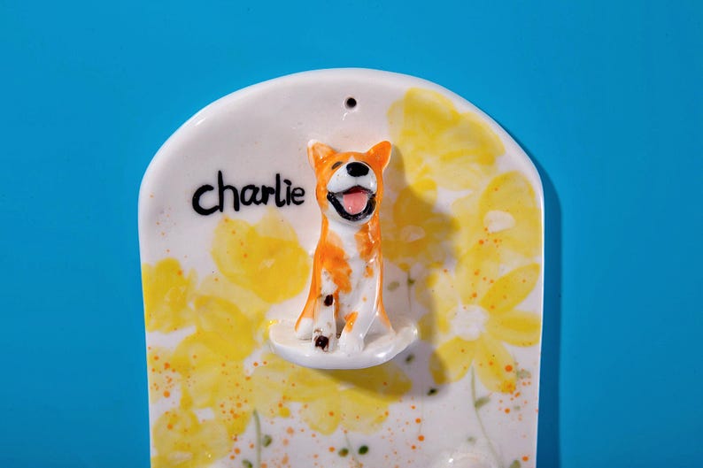 May include: A ceramic wall hanging featuring a smiling orange and white dog figurine. The dog is perched on a white base, with the name "Charlie" written above. The background is decorated with yellow floral patterns.