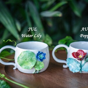 May include: Two white ceramic mugs with floral designs. One mug features a water lily design with green leaves and a blue flower, labeled "JUL Water Lily." The other mug has a red poppy design, labeled "AUG Poppy."