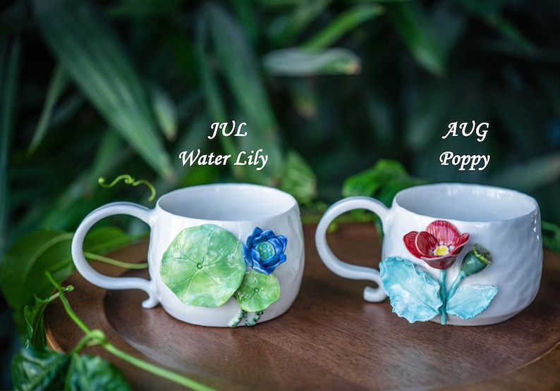 May include: Two white ceramic mugs with floral designs. One mug features a water lily design with green leaves and a blue flower, labeled "JUL Water Lily." The other mug has a red poppy design, labeled "AUG Poppy."