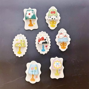 May include: A collection of seven decorative ceramic lamp ornaments. Each lamp features a unique design, including floral and fruit motifs, with a variety of colors such as green, pink, and blue. The lamps are mounted on white, scalloped-edge plaques.