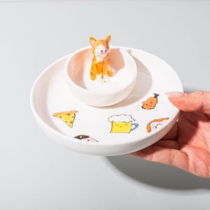 May include: A white ceramic serving dish with a central bowl, adorned with a cartoon cat figurine. The dish is decorated with colourful food illustrations, including pizza and beer. A hand is holding the dish.