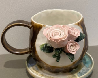 May include: A brown ceramic mug with a white interior and a 3D floral design featuring pink roses. The mug has a handle and is sitting on a matching saucer with a blue and white glaze.