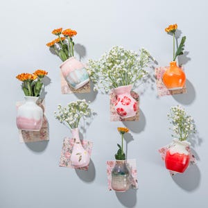 Könnte beinhalten: Eine Sammlung kleiner, farbenfroher Keramikvasen, jede mit frischen Blumen. Die Vasen haben verschiedene Formen und Farben, darunter Rosa, Orange und Blau, und sind auf dekorativem Papier befestigt. Die Blumen umfassen orangefarbene Chrysanthemen und weißen Schleierkraut.