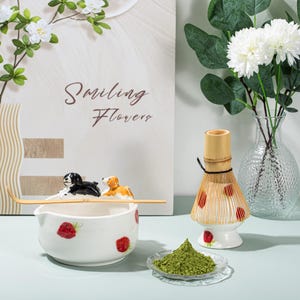 May include: A matcha tea set featuring a white ceramic bowl with strawberry designs and a bamboo whisk. The bowl has a decorative bamboo spoon and two dog figurines. A small plate holds green matcha powder, and a bamboo whisk is also present.