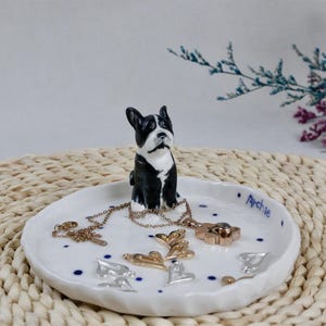 May include: A white ceramic trinket dish with a black and white Boston Terrier figurine. The dish holds necklaces and heart-shaped charms. The name "Archie" is written on the dish in blue.