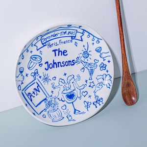 May include: White ceramic plate with blue hand-drawn illustrations. The plate features the text "The Johnsons", the date "September 17th 2025", and the location "Paris, France". Decorative elements include hearts, flowers, and champagne glasses.