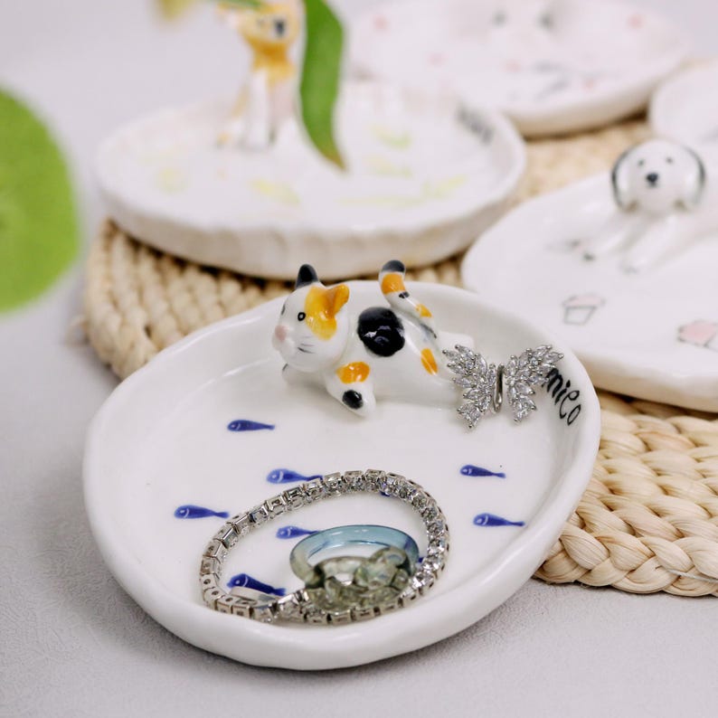 Puede incluir: Plato de cer&aacute;mica blanca para joyas con una peque&ntilde;a figura de gato, dise&ntilde;os de peces azules y un dije de mariposa plateado. El plato est&aacute; lleno de anillos plateados y una pulsera.