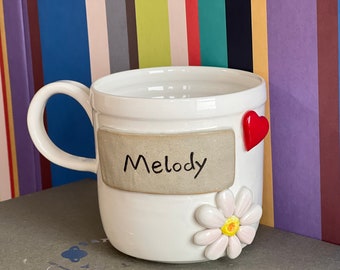 May include: White ceramic mug with a brown label that says "Melody". The mug has a red heart and a white daisy with a yellow center.