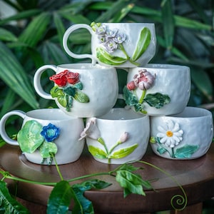 May include: A stack of six white ceramic mugs, each adorned with a different floral design. The flowers include roses, daisies, and other blooms in shades of red, pink, blue, and white. The mugs are arranged on a wooden surface against a backdrop of green foliage.