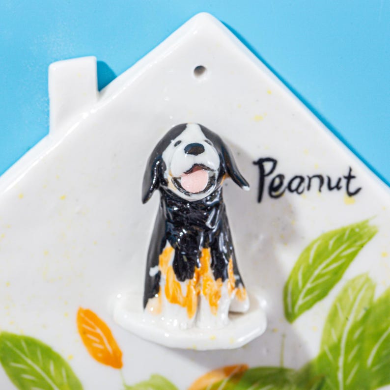 May include: A white ceramic house-shaped ornament with a black, white, and orange dog figurine. The word "Peanut" is written in black script. Green and orange leaf details are also present.