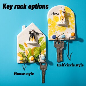 May include: Two ceramic key racks in house and half-circle styles, each with a dog figurine. The house-shaped rack has the name "Peanut" and the half-circle rack has the name "Charlie". The text "Key rack options" is at the top.