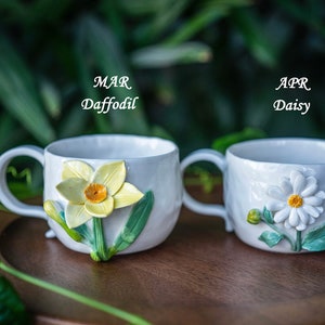 May include: Two white ceramic mugs with floral designs. One mug features a daffodil with yellow petals and an orange center, the other a daisy with white petals and a yellow center. The text "MAR Daffodil" and "APR Daisy" are visible.
