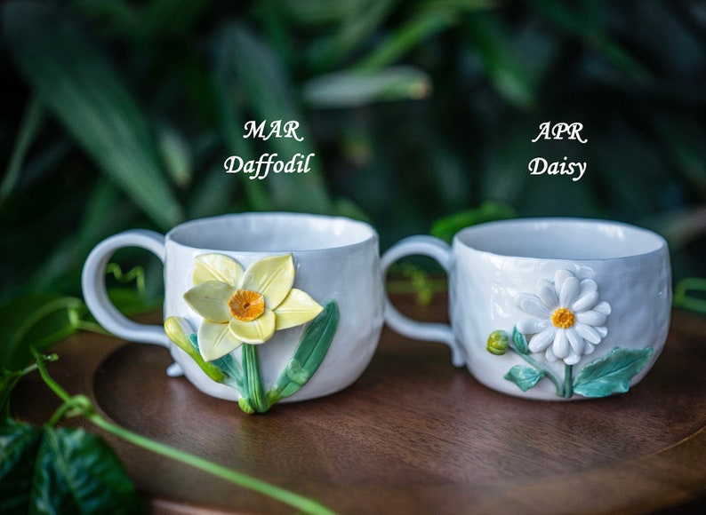 May include: Two white ceramic mugs with floral designs. One mug features a daffodil with yellow petals and an orange center, the other a daisy with white petals and a yellow center. The text "MAR Daffodil" and "APR Daisy" are visible.