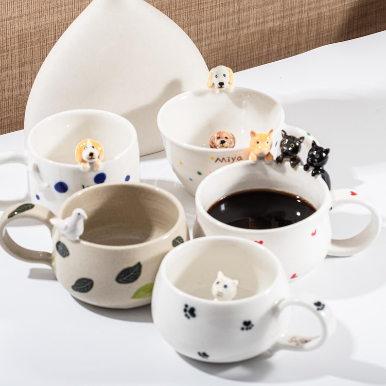 May include: A collection of ceramic mugs with animal figurines. The mugs feature dog, cat, and bird designs. Some mugs have blue polka dots, green leaves, and paw prints. One mug is filled with dark liquid.