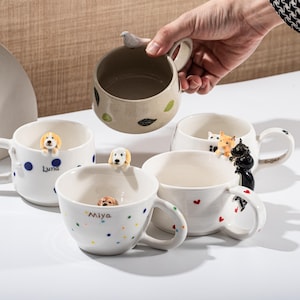 May include: A collection of ceramic mugs with unique designs. One mug is beige with leaf accents and a bird handle. Other mugs are white with dog and cat figurines, polka dots, and heart details. The mugs are of various sizes.