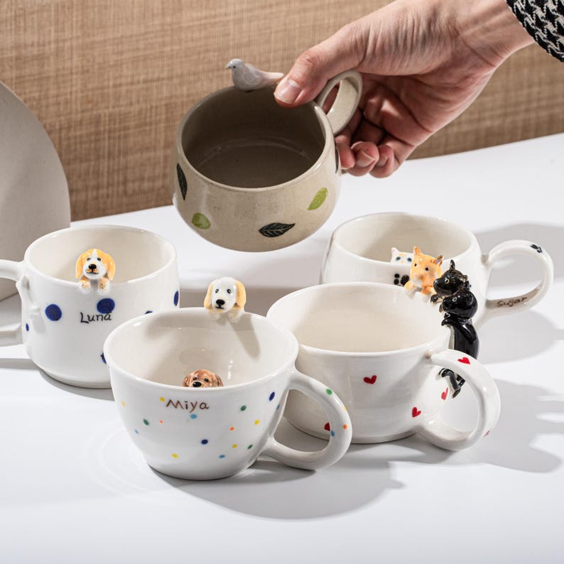 May include: A collection of ceramic mugs with unique designs. One mug is beige with leaf accents and a bird handle. Other mugs are white with dog and cat figurines, polka dots, and heart details. The mugs are of various sizes.