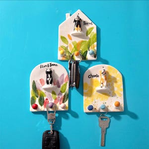 May include: Three ceramic key holders with pet-themed designs. One is house-shaped with a dog figurine and the name "Peanut". Another has a cat figurine and the names "Liam & Emma". The third has a dog figurine and the name "Charlie".
