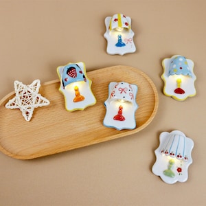 May include: Five ceramic nightlight lamps in various colours and designs. Each lamp features a small light bulb and a decorative lampshade. The lamps are arranged on a wooden surface.