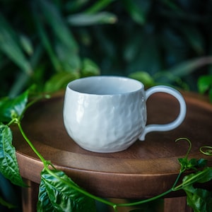 May include: A white ceramic mug with a textured surface and a curved handle. The mug sits on a wooden surface, surrounded by green foliage. The mug's design is simple and elegant, suitable for coffee or tea.