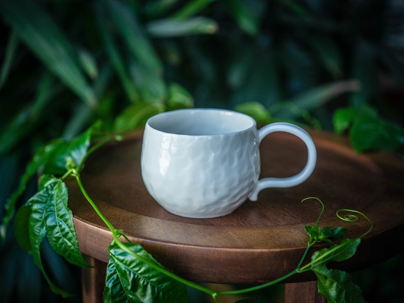 May include: A white ceramic mug with a textured surface and a curved handle. The mug sits on a wooden surface, surrounded by green foliage. The mug's design is simple and elegant, suitable for coffee or tea.
