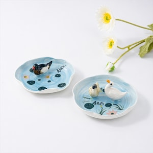 May include: Two ceramic decorative plates with a water lily design. One plate features a duck, the other has two ducks. The plates are painted with blue and white colours, with the names "Alex" and "A&B" on them.