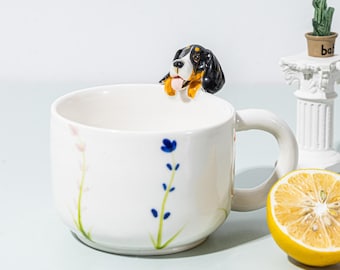 Puede incluir: Taza de cerámica blanca con una cabeza de perro decorativa en el borde. La taza presenta diseños florales pintados a mano en azul y rosa. También se ve un medio limón y un pequeño cactus en una maceta.