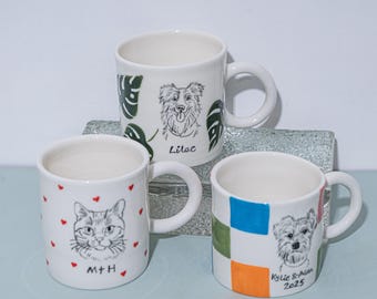Może przedstawiać: Trzy białe ceramiczne kubki z unikalnymi wzorami. Jeden kubek przedstawia ilustrację kota z czerwonymi serduszkami i inicjałami "M+H". Drugi ma portret psa i imię "Lilac". Trzeci kubek ma portret psa i imiona "Kylie & Alan 2025".