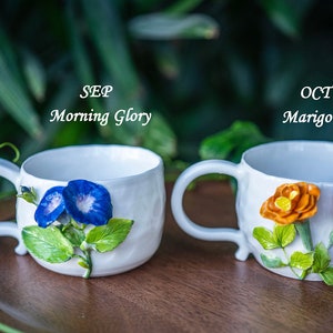May include: Two white ceramic mugs with floral designs. One mug features blue Morning Glory flowers, the other has orange Marigold flowers. The mugs have curved handles and are set on a wooden surface. Text on the mugs reads "SEP Morning Glory" and "OCT Marigold".