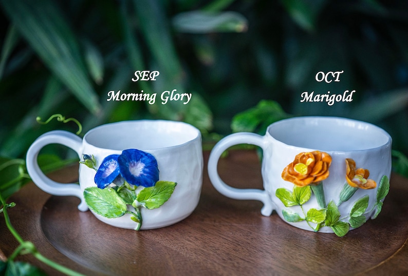 May include: Two white ceramic mugs with floral designs. One mug features blue Morning Glory flowers, the other has orange Marigold flowers. The mugs have curved handles and are set on a wooden surface. Text on the mugs reads "SEP Morning Glory" and "OCT Marigold".