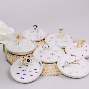 Puede incluir: Conjunto de ocho platos de cerámica blanca para joyas con figuritas de animales. Cada plato presenta un animal diferente, incluyendo un perro, un gato, un pájaro y una ardilla. Los platos están decorados con varios patrones, incluyendo huellas de patas, huesos y peces.