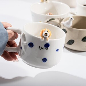 May include: White ceramic mug with blue polka dots and a small, orange and white dog figurine on the rim. The mug has the name "Luna" printed on the side. Other mugs with animal figurines are visible in the background.