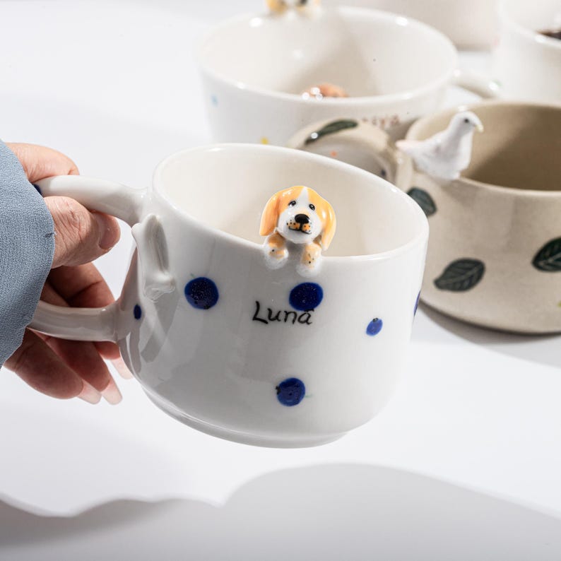 May include: White ceramic mug with blue polka dots and a small, orange and white dog figurine on the rim. The mug has the name "Luna" printed on the side. Other mugs with animal figurines are visible in the background.