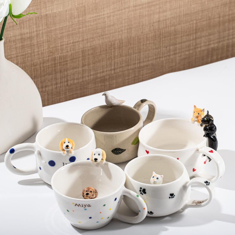 May include: A collection of ceramic mugs with animal-themed designs. The mugs feature 3D dog, cat, and bird figurines. Some mugs have polka dots, paw prints, and heart designs. One mug has the name "Miya" on it.