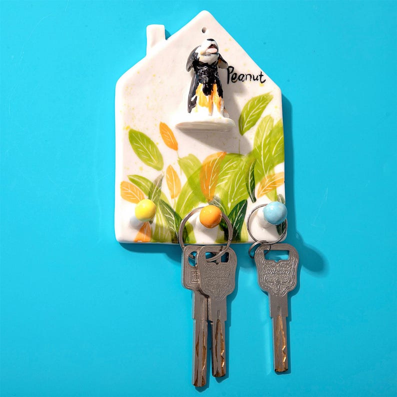 May include: A white ceramic house-shaped key holder with a dog figurine and the name "Peanut". The holder features green and orange leaf and fruit designs, with three key rings holding silver keys. The background is a solid blue.