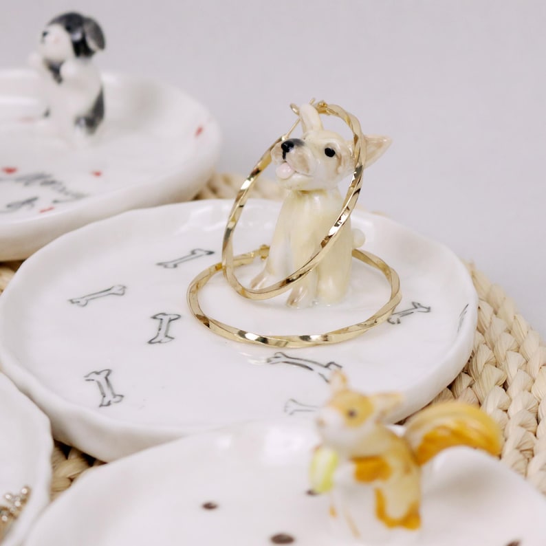 Puede incluir: Plato de cer&aacute;mica blanco para anillos con una peque&ntilde;a figura de perro sosteniendo un par de aros de oro. El plato est&aacute; decorado con dise&ntilde;os de huesos negros.