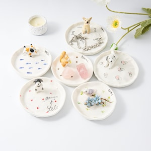 Puede incluir: Seis pequeñas bandejas de cerámica blanca con diferentes figuritas de animales y diseños. Las bandejas están etiquetadas con nombres como "Milo", "Peanut", "Sally" y "Morning & Lila".