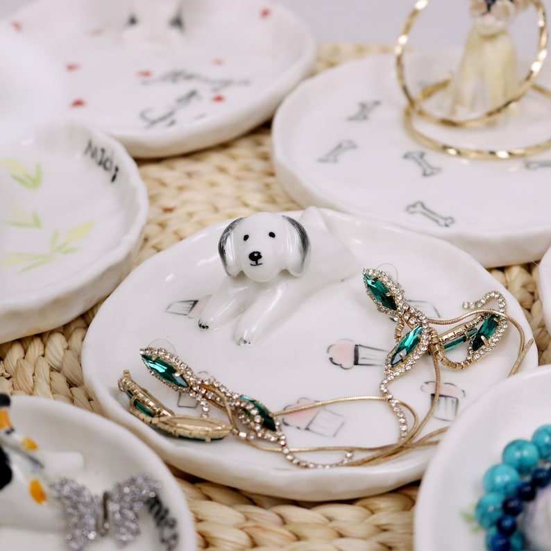 Puede incluir: Plato de anillo de cer&aacute;mica blanca con una peque&ntilde;a figurita de perro blanco, pendientes de cristal verde y transparente y un collar de cadena dorada. El plato tiene un fondo blanco con un contorno negro de una magdalena y un hueso.