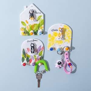 Op de afbeelding: Vier keramische sleutelhangers met honden- en kattenbeeldjes. De sleutelhangers zijn wit met kleurrijke accenten en de namen "Peanut", "Charlie" en "Liam & Emma". Eén sleutelhanger heeft een groene sleutelring en een sleutel.