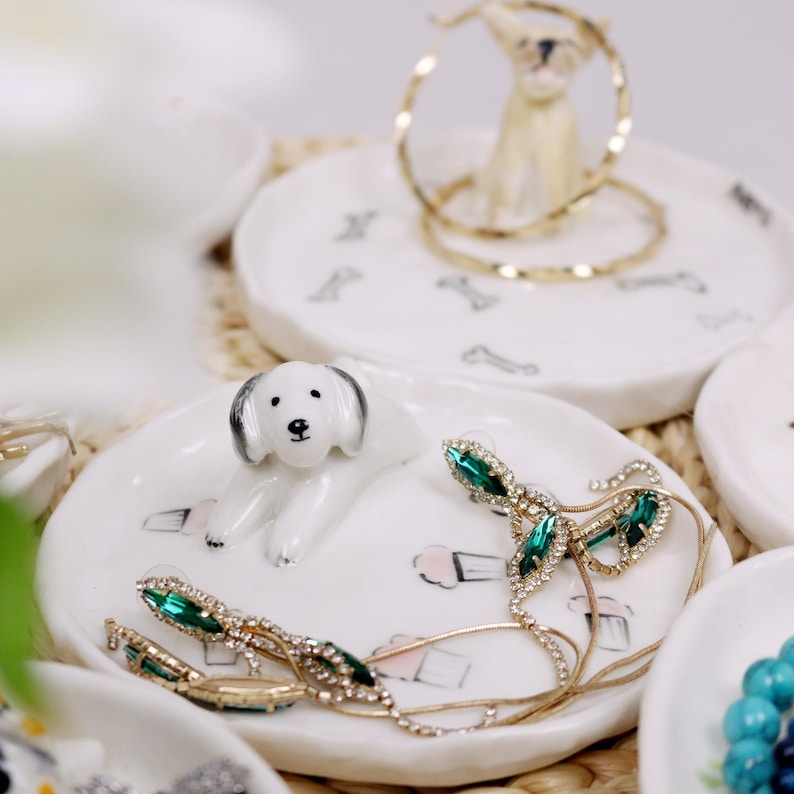 Puede incluir: Plato de cer&aacute;mica blanco para anillos con una peque&ntilde;a figura de perro blanco, pendientes de piedras preciosas verdes y un collar de cadena dorada. El plato tiene un patr&oacute;n de peque&ntilde;os conos de helado.