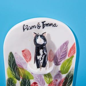 May include: A ceramic cat figurine with black and white markings, perched on a white base. The figurine is surrounded by colorful leaf designs in red, green, and purple. The names "Liam & Emma" are written above the cat.