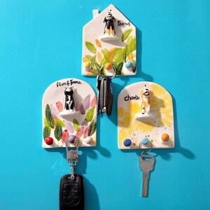 May include: Three ceramic key holders with dog figurines. The key holders are white with colorful leaf and floral designs. Each key holder has a different dog figurine and name: Peanut, Liam & Emma, and Charlie. The key holders are mounted on a blue wall.