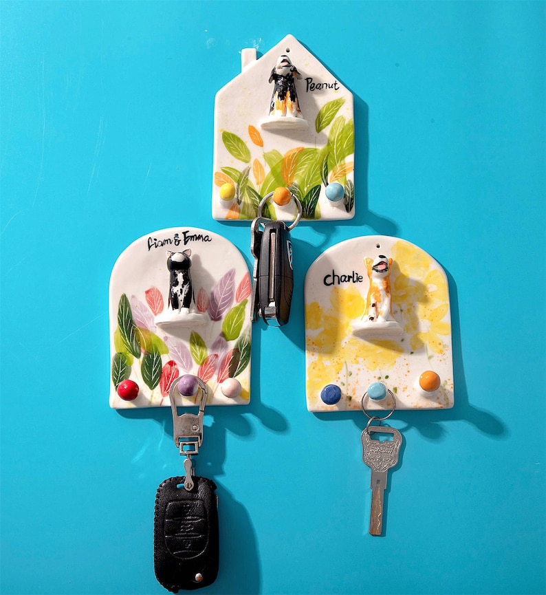 May include: Three ceramic key holders with dog figurines. The key holders are white with colorful leaf and floral designs. Each key holder has a different dog figurine and name: Peanut, Liam & Emma, and Charlie. The key holders are mounted on a blue wall.