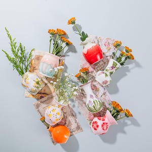 Könnte beinhalten: Eine Sammlung kleiner, farbenfroher Keramikvasen, jede mit frischen orangefarbenen Blumen und Grün. Die Vasen weisen verschiedene Designs auf, darunter Blumenmuster und unifarbene Farben. Einige Vasen sind rosa, rot und orange. Die Anordnung befindet sich vor einem hellblauen Hintergrund.