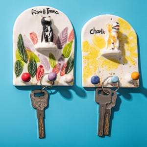 May include: Two ceramic key holders with pet-themed designs. One features a black and white cat, the other a yellow and white dog. Each has a name above the figurine and colorful leaf or floral accents, with key hooks below.