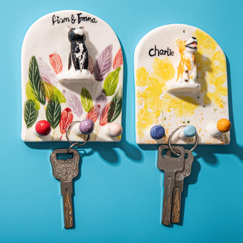 May include: Two ceramic key holders with pet-themed designs. One features a black and white cat, the other a yellow and white dog. Each has a name above the figurine and colorful leaf or floral accents, with key hooks below.