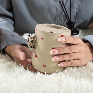 May include: A beige ceramic mug adorned with small red hearts and a 3D figurine of a dog. The mug has a small opening near the top and is held by a person. The mug is on a white fluffy surface.