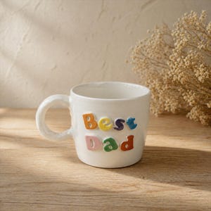 May include: White ceramic mug with the words "Best Dad" in colorful, raised letters. The mug has a rounded handle and sits on a wooden surface. The letters are in orange, yellow, blue, green, and red.